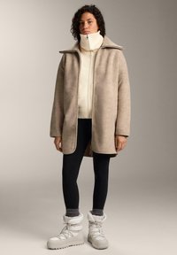 Beige fleecetakki leveällä kauluksella, päällä valkoinen vetoketjullinen neule, yhdistetty mustiin leggingsseihin ja valkoisiin talvisaappaisiin, joissa on turkisreunus.