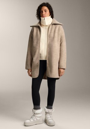 Béžový flísový kabát so širokým golierom, nosený cez biely sveter na zips, v kombinácii s čiernymi legínami a bielymi zimnými čižmami s kožušinovou úpravou.