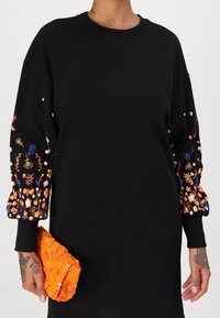 Svart sweatshirtklänning med broderade blommor i orange, rosa och blått på ärmarna; håller en texturerad orange kuvertväska.