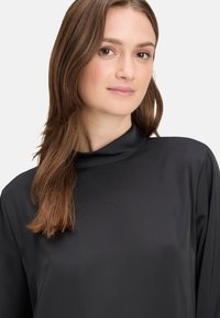Schwarze Bluse mit hohem Kragen aus glattem, leichtem Stoff. Sie hat lange Ärmel und einen lockeren Schnitt mit minimalen Nähten für einen klaren Look.