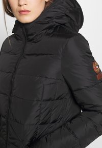 Black puffer jacket with a shiny finish, quilted design, high collar, and a zip closure; features a round logo patch on the sleeve.