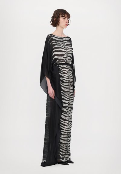 Femeie îmbrăcată într-o rochie lungă cu dungi zebra alb-negru, având un detaliu dintr-o capa neagră transparentă care flutură, stând în fața unui fundal alb simplu.