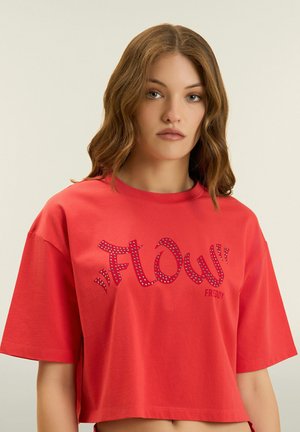 Joven con cabello castaño claro ondulado que lleva una camiseta roja recortada con el texto "FLOW" decorado con pequeños tachuelas.
