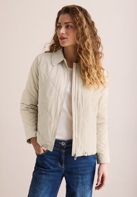 Veste matelassée beige clair avec fermeture éclair et col montant, comportant deux poches latérales et des détails de texture subtils. Portée sur un top blanc.