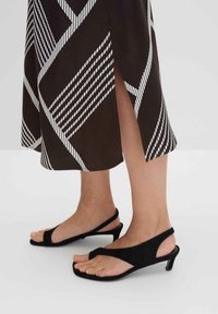Sandali con tacco in suede nero con design a toe post e una sottile cinghia attorno alla caviglia, abbinati a una gonna marrone con motivi diagonali bianchi.