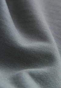 Pelēka auduma ar gludu tekstūru, kuram ir smalks austs raksts. Materiāls izskatās viegls un elastīgs, maigi krītot.