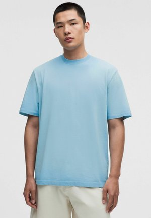 Junger Mann steht und trägt ein schlichtes hellblaues oversized T-Shirt mit kurzen Ärmeln und hellbeigen Hosen vor einem einfarbigen Hintergrund.