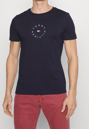 Mężczyzna w granatowej koszulce Tommy Hilfiger z okrągłym logo i czerwonych spodniach, stoi na tle jasnego, jednolitego tła.