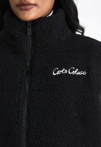 Crna flis jakna s visokim ovratnikom, s prednjim zatvaračem i bijelim izvezenim logotipom "Carlo Colucci." Mekana tekstura i udoban dizajn.