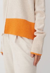 Beżowy sweter dzianinowy z pomarańczowym ściągaczem u dołu i na mankietach. Sweter ma luźny krój i miękką strukturę, zestawiony z kremowymi spodniami.