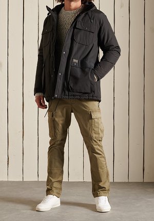 Personne portant une veste noire à capuche, un pull en tricot gris, un pantalon cargo beige et des baskets blanches, debout contre un mur en bois blanc.