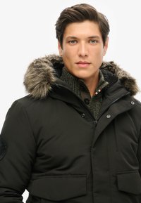 Parka vert foncé avec capuche en fourrure, fermeture éclair et poche poitrine. Superposée à un pull tricoté avec un motif texturé.