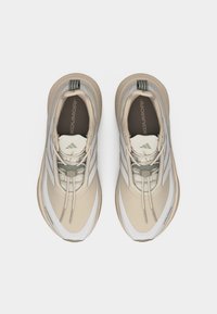 Pereche de pantofi atletici bej și albi cu șireturi, cu mânere gri și tălpi interioare marcate, văzuți de sus pe un fundal alb.