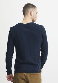 Tamno plava košulja dugih rukava od glatkog materijala, sa okruglim izrezom i uskim manžetama, nosi se uz khaki hlače.