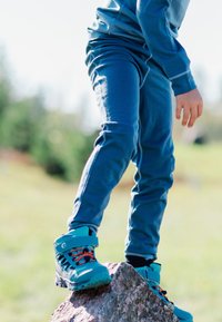 Blå sportsbukser parret med teal vandrestøvler med orange snørebånd, stående på en sten i et græsset udendørs miljø.