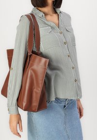 Donna che indossa una camicia a righe verdi e bianche con bottoni e una gonna di jeans azzurro chiaro, con una borsa a tracolla in pelle marrone.