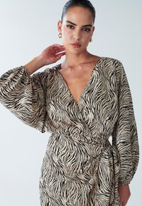 Dierlijke print jurk in beige en zwart, met een diepe V-hals, wijde mouwen en een gestrikt middel. Glad materiaal met een vloeiende textuur.