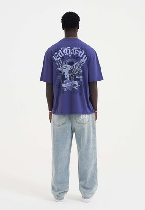 Purple short-sleeve t-shirt featuring a graphic design on the back; paired with light blue, relaxed-fit jeans.