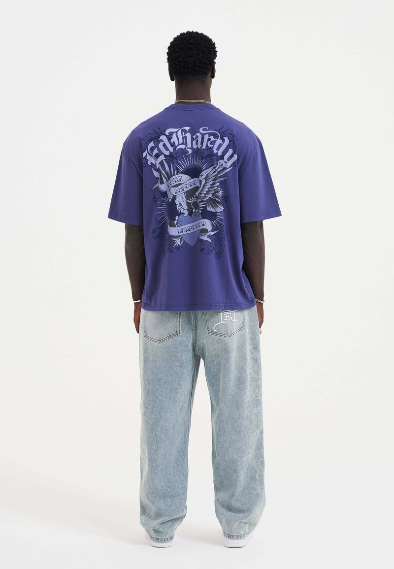 Paarse T-shirt met korte mouwen met een grafisch ontwerp op de achterkant; gecombineerd met lichtblauwe, relaxte jeans.
