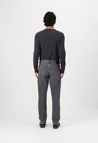 Hombre con pelo rizado viste un suéter gris oscuro, jeans grises y zapatos negros, de pie frente a un fondo blanco con las manos relajadas a los lados.