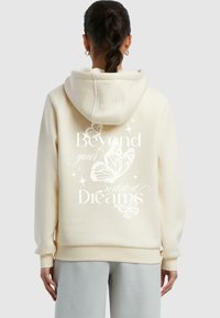 Kremfarget hettegenser med avslappet passform, med et sommerfugl-design og teksten "Beyond your wildest Dreams" i hvitt på ryggen.