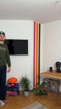 Τηλεόραση τοποθετημένη σε τοίχο με πορτοκαλί και ριγέ ροζ διακοσμητικά στοιχεία στους τοίχους. Ένα μπλε τραπεζάκι με πορτοκαλί λάμπα, γλαστράκια φυτών και ένα ξύλινο γραφείο.