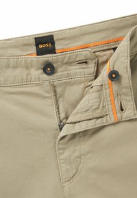 Pantalones beige con cierre de cremallera y botón, que presentan un acento naranja en la cintura. La tela parece suave con textura mínima.