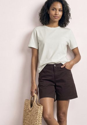 Femme portant un t-shirt blanc et un short foncé tenant un sac fourre-tout en paille tressée, debout devant un mur rose clair.