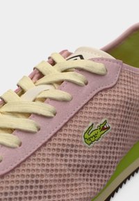 Ροζ αθλητικό παπούτσι με πλέγμα, κίτρινες κορδόνια, λευκή γλώσσα, πράσινη επένδυση και πράσινο λογότυπο κροκόδειλου. Υπέροχη υφή στο πάνω μέρος και αντίθετη σόλα.