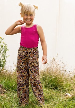 Roze geribbelde tanktop gecombineerd met wijduitlopende broeken met een paisleypatroon in goud, paars en bruin. Model staat in een grasveld en glimlacht.