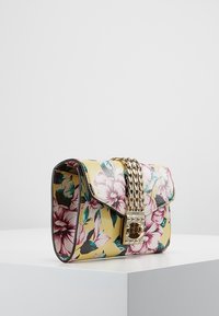 Gul blomsterhandtaske med pink og grønne blomster, guld kædestropp, og guld dreje-lås, hvilende på hvid overflade.