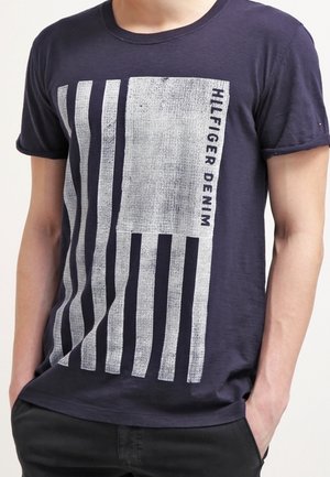 Marineblaues Baumwoll-T-Shirt mit einem silbernen Grafikdesign aus vertikalen Streifen und einem rechteckigen Block, mit "HILFIGER DENIM" vertikal gedruckt.