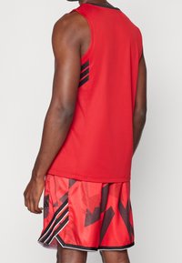 Débardeur de sport rouge avec des accents noirs, associé à un short rouge présentant des motifs abstraits noirs et des bords rayés. Texture de tissu lisse.