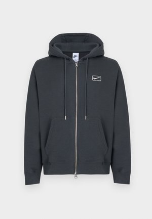 Sweat à capuche zippé gris foncé avec poches avant, capuche ajustable avec cordon de serrage, et petit logo Nike blanc sur le côté gauche de la poitrine.