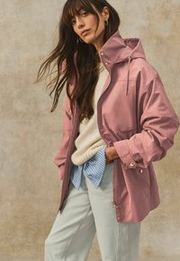 Rosa wasserdichte Jacke mit Kapuze, glänzende Oberfläche, Reißverschluss vorne; über einem cremefarbenen Pullover und einem blau gestreiften Hemd getragen, kombiniert mit hellen Jeans.