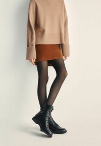 Osoba ubrana w brązową spódnicę, oversize'owy beżowy sweter, przezroczyste czarne rajstopy i czarne sznurowane botki do kostek stoi na jednolitym tle.