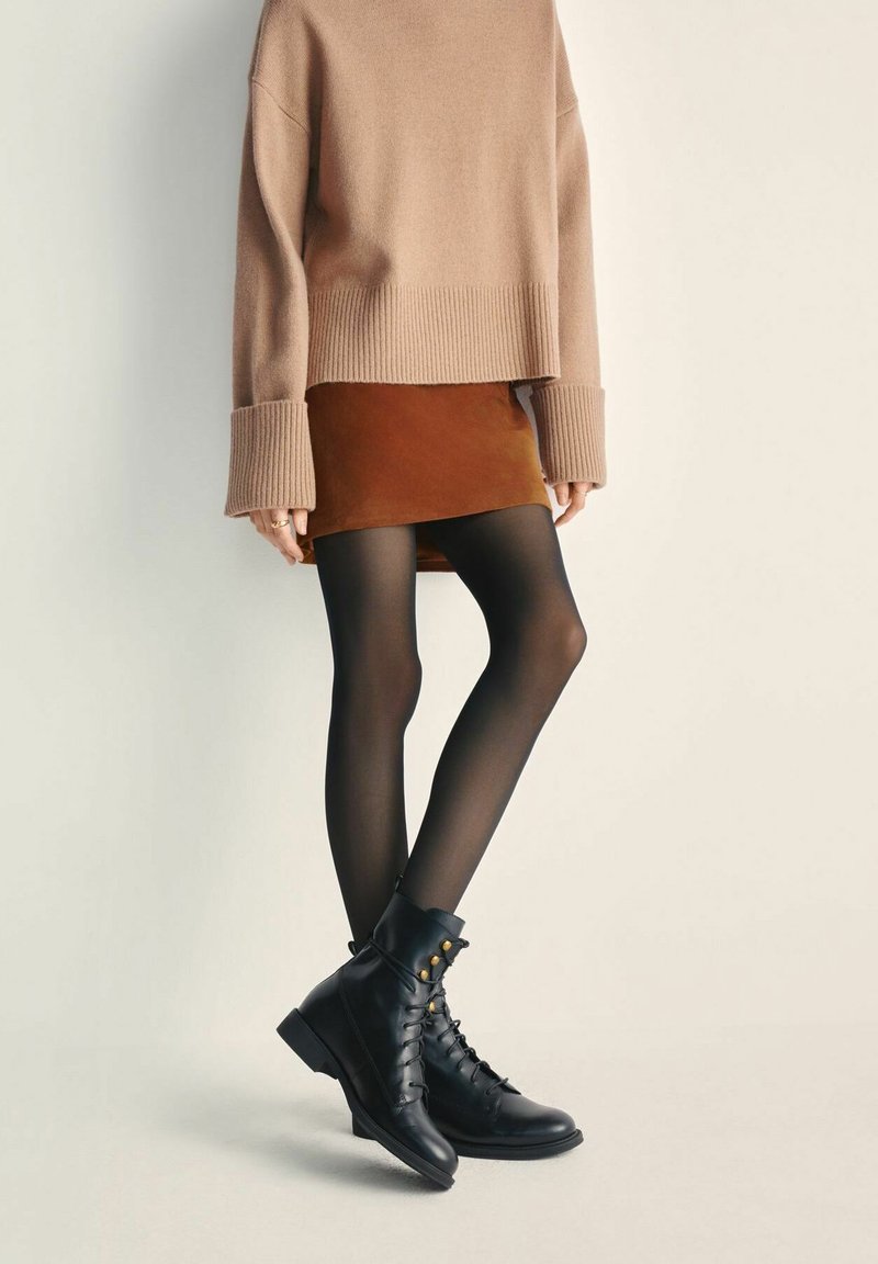 Osoba ubrana w brązową spódnicę, oversize'owy beżowy sweter, przezroczyste czarne rajstopy i czarne sznurowane botki do kostek stoi na jednolitym tle.