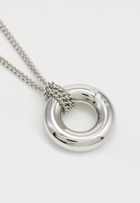 Pendentif argenté circulaire sur une chaîne, présentant une surface métallique polie avec une petite pierre claire et un détail de chaîne nouée.