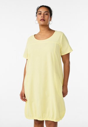 Femme portant une robe ample jaune pâle à manches courtes, avec un col arrondi et un ourlet élastique, debout devant un fond blanc.