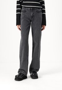Jeans acampanados de cintura alta en denim gris oscuro, con cinco bolsillos y costuras sutiles. Combinados con zapatos de plataforma negros gruesos.