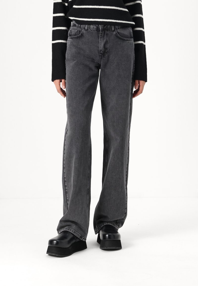 Jeans acampanados de cintura alta en denim gris oscuro, con cinco bolsillos y costuras sutiles. Combinados con zapatos de plataforma negros gruesos.