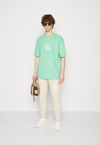 Camiseta de algodón verde con un logo blanco, combinada con pantalones jogger crema que presentan un detalle del logo. El modelo sostiene un pequeño bolso marrón y lleva puestas gafas de sol.