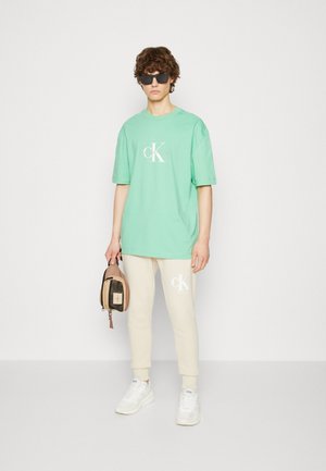T-shirt in cotone verde con logo bianco, abbinato a pantaloni joggers beige con dettaglio del logo. Il modello tiene una piccola borsa marrone e indossa occhiali da sole.