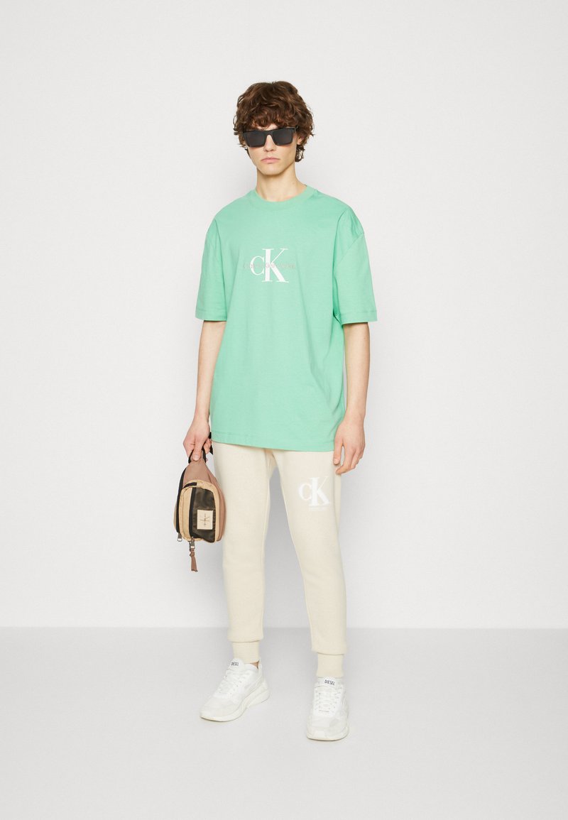Camiseta de algodón verde con un logo blanco, combinada con pantalones jogger crema que presentan un detalle del logo. El modelo sostiene un pequeño bolso marrón y lleva puestas gafas de sol.
