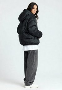 Doudoune noire avec capuche, à motif matelassé, ourlet élastique, portée sur une chemise blanche et associée à un pantalon large gris et des baskets blanches.