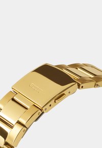 Montre en métal doré avec une finition polie, présentant un large bracelet avec des maillons reliés et un fermoir rectangulaire.