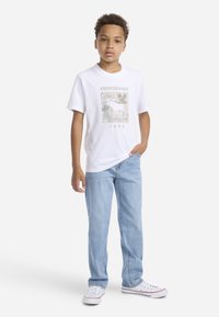 Biała koszulka z krótkim rękawem z graficznym nadrukiem przedstawiającym zwierzę z rogami oraz napisem "Abercrombie 1892", zestawiona z jasnoniebieskimi dżinsami.