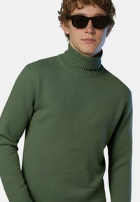 Maglione verde a collo alto con texture a coste e maniche lunghe. Il colletto è alto e il tessuto appare liscio e aderente. Indossato con occhiali da sole scuri.