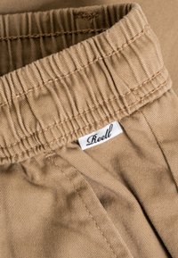 Primer plano de la cintura elástica beige en los pantalones con una etiqueta de tela blanca de la marca cosida por dentro que dice "Reell".