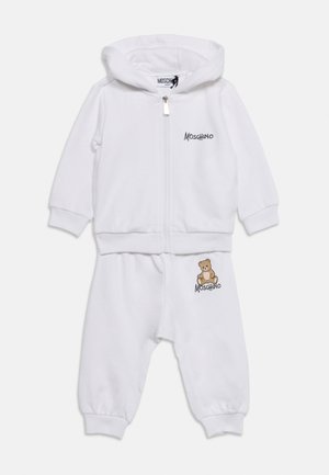 Biały dziecięcy dres z kapturem i zamkiem błyskawicznym, tekst ""Moschino"" na piersi i spodniach oraz grafika misia na spodniach.